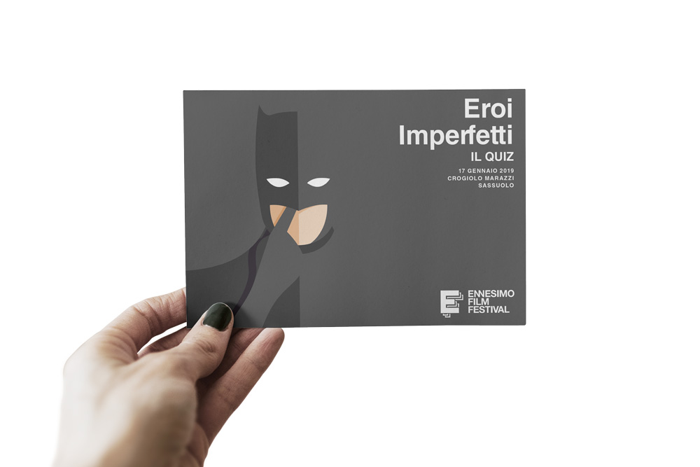 Eroi Imperfetti: la cartolina in formato finale.
Una mano regge una cartolina usata durante una delle serate delle proiezioni. In particolare, in questa occasione si vede chiaramente Batman con il dito nel naso, il nome della serata e il logo di Ennesimo Film Festival.